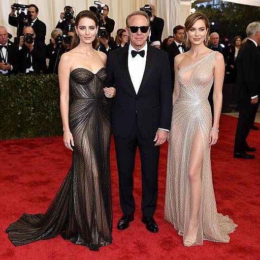 Photograph of three people on a red carpet: an older man in a black tuxedo with a bow tie, and two women in glittering