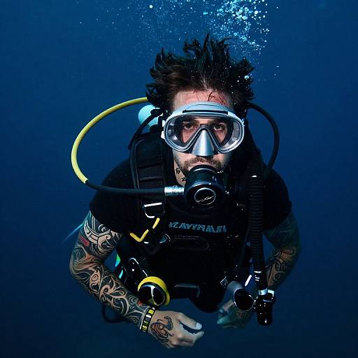 Photograph of a tattooed male scuba diver with wild hair, wearing a mask, regulator, and yellow hose, underwater in deep blue.