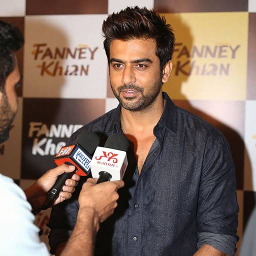 Photograph of a handsome, dark-haired man with a trimmed beard in a navy blue shirt, being interviewed by a reporter holding a 