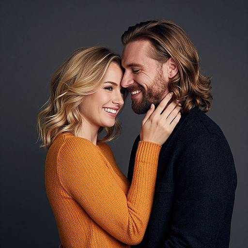 Photograph of a smiling couple, woman with blonde wavy hair in an orange sweater, man with brown hair and beard in a black sweater, touching