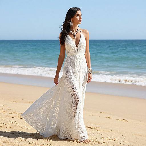 Photograph of a dark-haired woman in a white lace wedding dress standing on a sunny beach with blue ocean and clear sky.