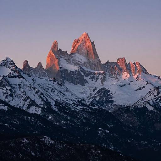 Photograph of a majestic, snow-capped mountain range at sunrise, with jagged peaks bathed in pink and orange light against a clear blue sky
