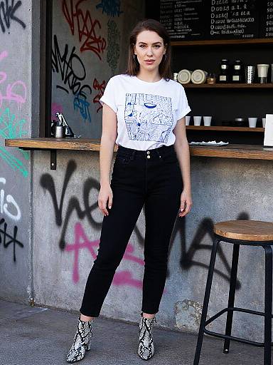 Photograph of a young woman with fair skin and brown hair, wearing a white graphic tee, black pants, and snake-print boots, standing in front
