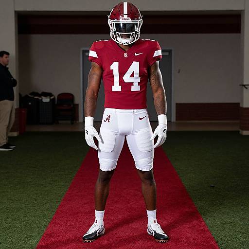 Photograph of an African-American male football player in red jersey number 14, white pants, and helmet, standing on green field with red carpet,