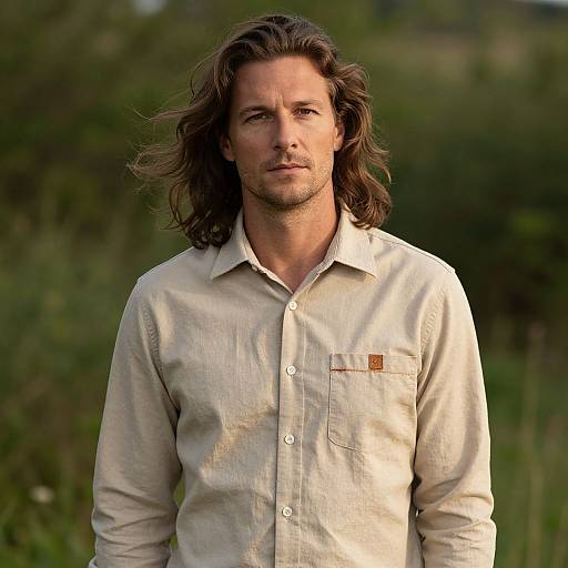 Photograph of a handsome man with long, wavy brown hair, wearing a beige button-up shirt, standing outdoors with a blurred green background.