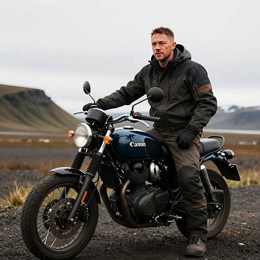 Photograph of a rugged, bearded man in black jacket and gloves, seated on a blue Canon motorcycle, in a rocky, mountainous landscape.
