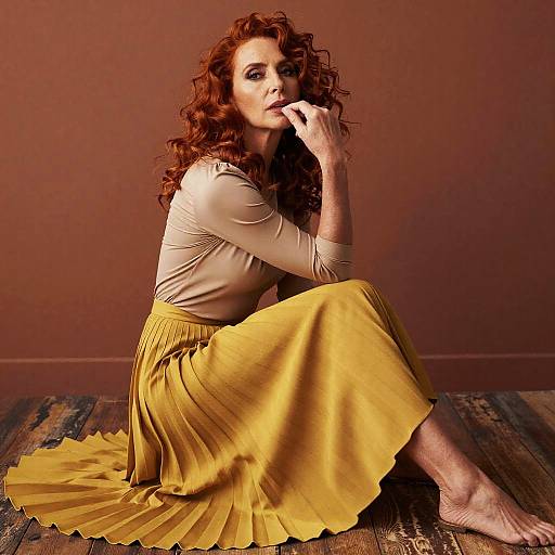 Photograph of a fair-skinned woman with curly red hair, wearing a beige top and yellow pleated skirt, sitting on a wooden floor against a