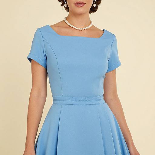 Photograph of a woman in a vintage-style, light blue, short-sleeved, A-line dress with a pearl necklace, standing against a plain