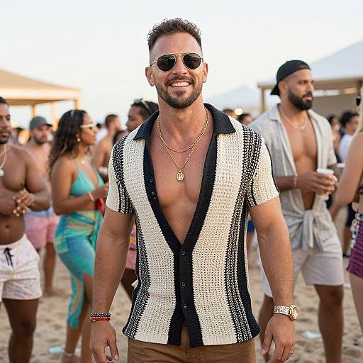 Photograph of a smiling, bearded man in a black and white patterned shirt, sunglasses, and necklace, standing on a busy beach in a