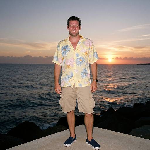 Photograph of a smiling man with short dark hair, wearing a floral shirt, beige shorts, and blue sneakers, standing on a rocky coastline at sunset