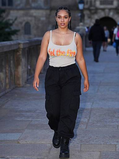Photograph of a confident woman with dark skin, braided hair, wearing a white tank top with orange flames, black cargo pants, and black boots