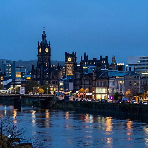 Photograph of a cityscape at dusk, featuring illuminated historic buildings with clock towers, reflected in a calm river, against a blue twilight sky.