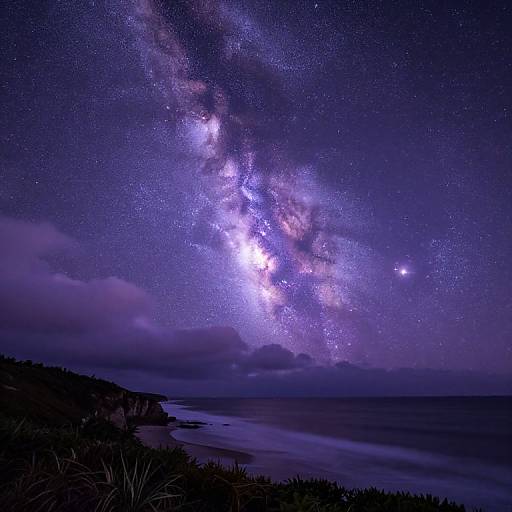 Photograph of a starry night sky with the Milky Way galaxy prominently visible, over a dark, cloudy coastline and calm sea.