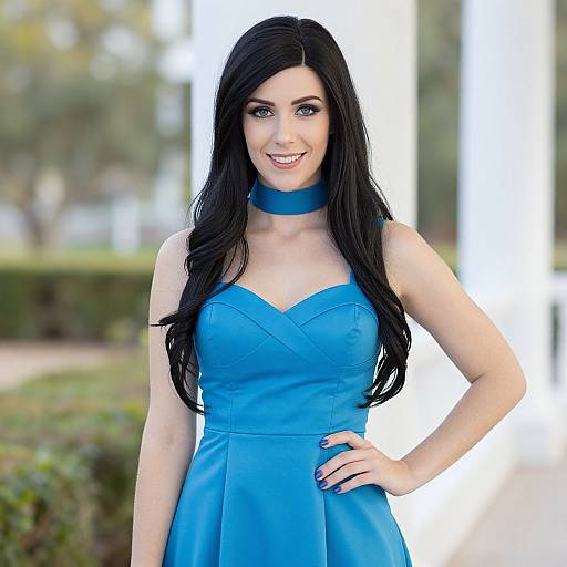 Photograph of a smiling woman with long black hair, blue strapless dress, and matching choker, standing outdoors with a blurred green and white background
