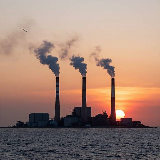 Photograph of a sunset over a silhouetted industrial plant with three tall smokestacks emitting dark smoke, standing against a vibrant orange and pink