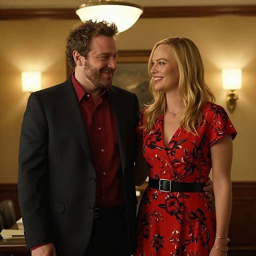 Photograph of a smiling, bearded man in a black suit and red shirt, standing next to a blonde woman in a red floral dress with a