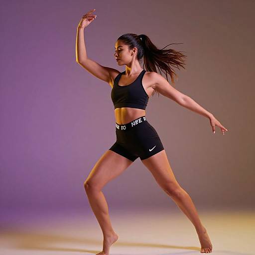 Photograph of a fit, tan-skinned woman in a black sports bra and shorts, mid-dance pose, with long dark hair, purple and