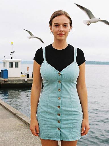Photograph of a fair-skinned woman with brown hair in a black t-shirt and light blue button-up dress, standing by a waterfront with seag
