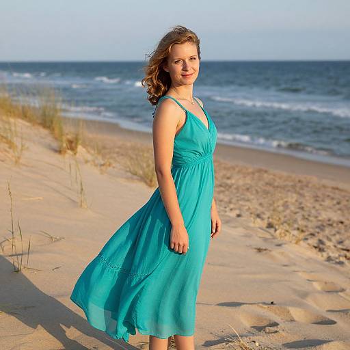 Photograph of a smiling, fair-skinned woman with curly brown hair, wearing a flowing turquoise dress, standing on a sandy beach with the ocean and