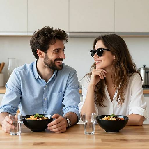 Photograph of a smiling couple with dark hair, wearing sunglasses and light blue and white shirts, eating salad at a wooden table in a bright, modern