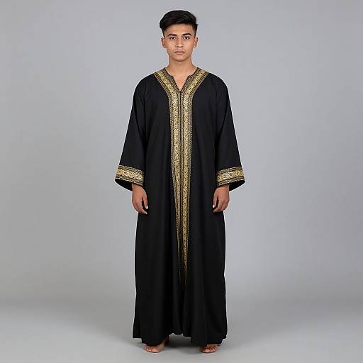 Photograph of a young South Asian man with short black hair, wearing a long black traditional tunic with gold embroidered trim, standing against a plain light