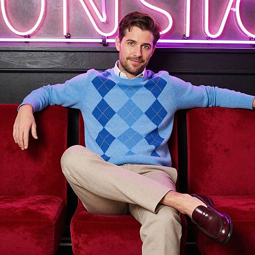 Photograph of a bearded man with dark hair, wearing a blue diamond-pattern sweater, beige pants, and brown shoes, sitting on red velvet chairs