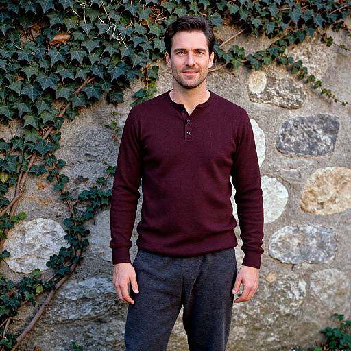 Photograph of a smiling, dark-haired man in a black long-sleeve henley and dark pants, standing against a stone wall covered in iv