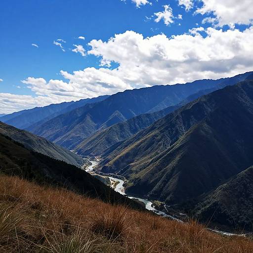 Photograph of a mountainous landscape with blue sky, scattered clouds, a winding river, and brown grass in the foreground.