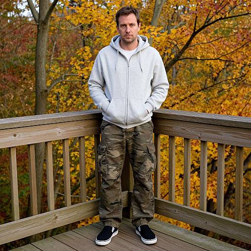 Photograph of a man with short brown hair, light beard, wearing a white hoodie, camouflage pants, and black sneakers, standing on a wooden deck