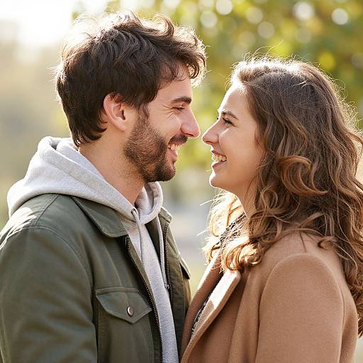 Photograph of a smiling couple facing each other outdoors, sunlight highlighting their faces; man in green jacket, white hoodie; woman in brown coat, w