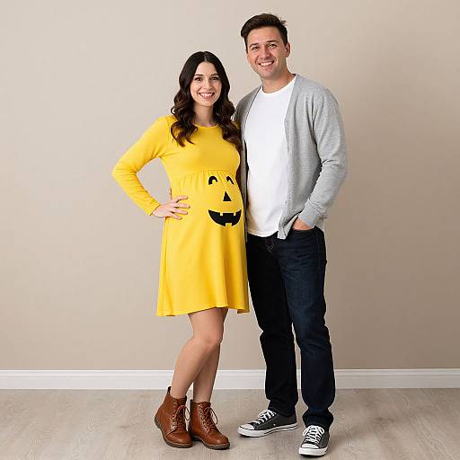 Photograph of a smiling couple; woman in yellow dress with black smiley face, brown boots, and long black hair; man in gray cardigan