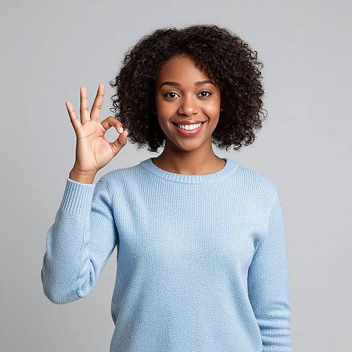Photograph of a smiling Black woman with curly hair, wearing a light blue sweater, holding up a peace sign with her right hand against a plain white
