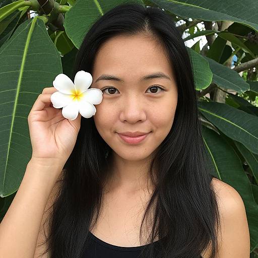 Photograph of an Asian woman with long black hair, holding a white plumeria flower against a backdrop of large green leaves.