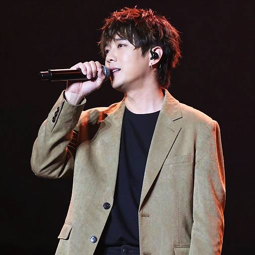 Photograph of a young Asian male singer with tousled black hair, wearing a beige blazer over a black shirt, holding a microphone, singing into