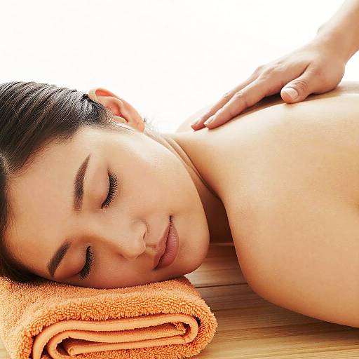 Photograph of a serene, light-skinned woman with dark hair, eyes closed, lying face down on an orange towel, receiving a massage on her