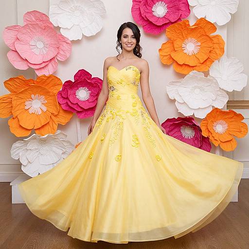 Photograph of a smiling woman in a strapless, yellow, floral-embroidered gown, standing in front of large, colorful paper flowers (
