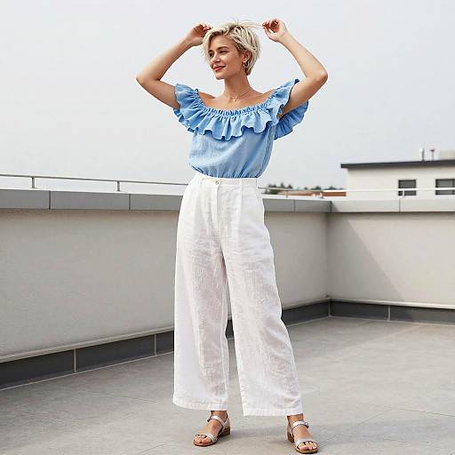 Photograph of a blonde woman with short hair, wearing a blue ruffle top and high-waisted white pants, standing on a rooftop with arms