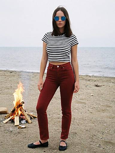 Photograph of a young woman with straight brown hair, wearing blue sunglasses, black-and-white striped crop top, red high-waisted jeans, and