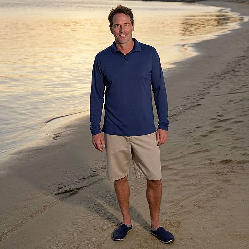 Photograph of a smiling middle-aged man with short brown hair, wearing a navy polo, beige shorts, and blue sneakers, standing on a sandy beach
