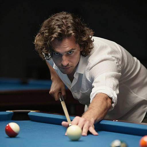 Photograph of a focused, curly-haired man in a white shirt, aiming a pool cue at a white ball on a blue pool table.