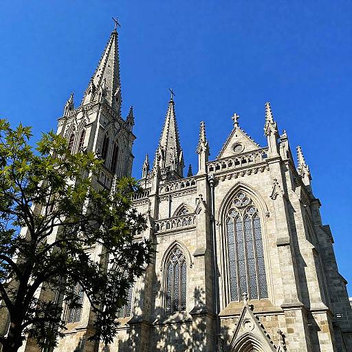 Photograph of a Gothic-style cathedral with two pointed spires, intricate stone carvings, tall arched windows, and clear blue sky, partially