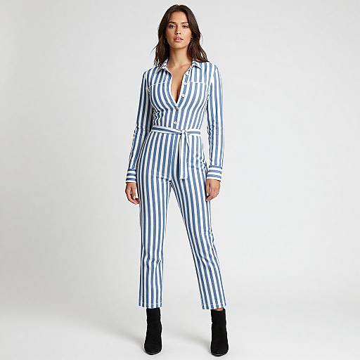 Photograph of a confident woman with straight brown hair, wearing a blue and white striped jumpsuit with a belted waist and black ankle boots, standing