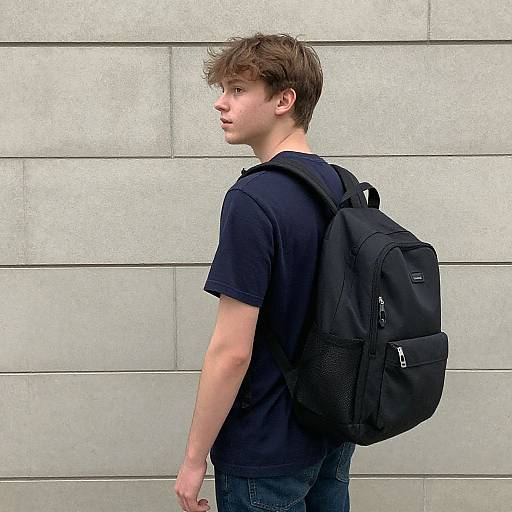 Photograph of a young Caucasian man with light brown hair, wearing a black t-shirt and blue jeans, carrying a black backpack, standing against a gray