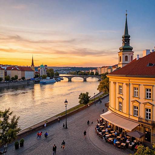 Photograph of a vibrant sunset over a European river city, featuring a historic building with a spire, cobblestone street, river, and people