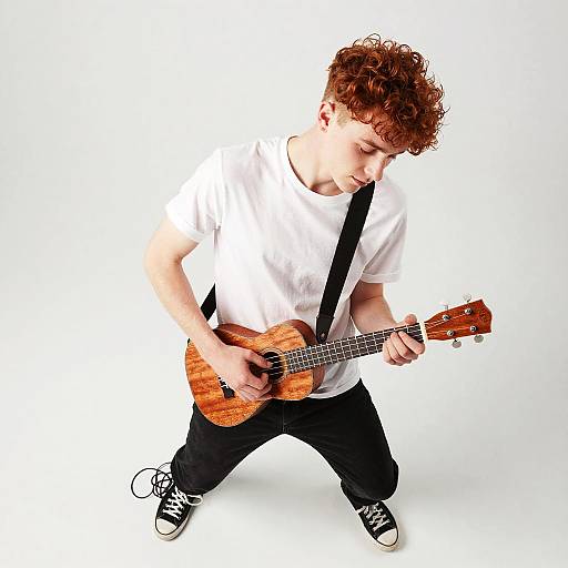 Photograph of a curly-haired young man playing a wooden acoustic guitar, wearing a white t-shirt, black pants, and black sneakers, against a white