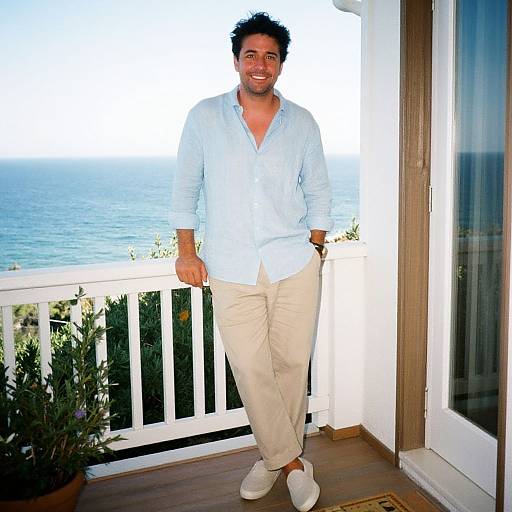 Photograph of a smiling, dark-haired man in a light blue shirt and beige pants, standing on a balcony with ocean view.