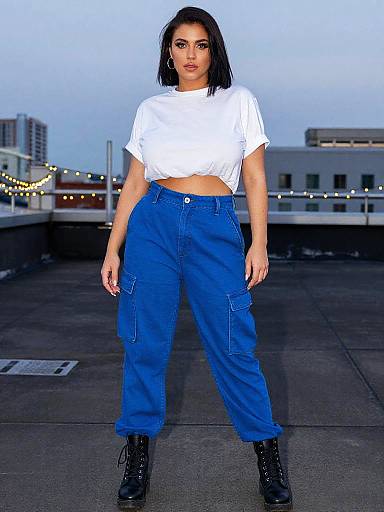 Photograph of a confident woman with dark hair, white crop top, blue high-waisted cargo pants, black boots, standing on a rooftop at