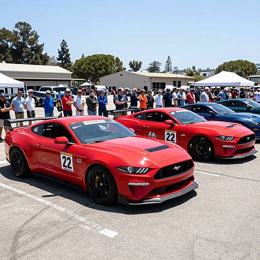 Photograph of two bright red Ford Mustangs with black wheels and number 22, parked in a sunny outdoor event with a crowd and white tents in