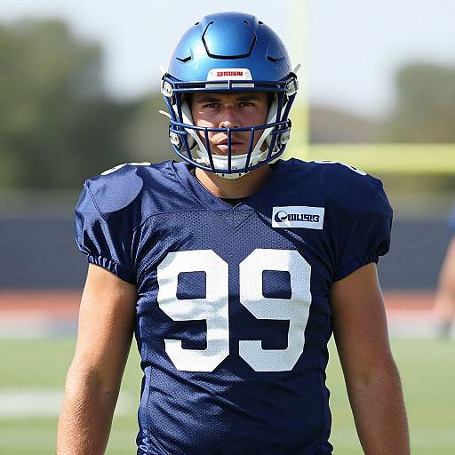 Photograph of a male football player in a navy blue helmet and jersey with the number 99, standing on a sunlit field.