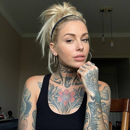 Photograph of a blonde woman with a ponytail, black tank top, extensive tattoos, nose ring, hoop earrings, and thinking pose in a dim